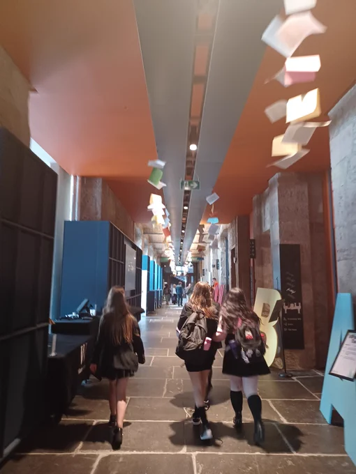 Image shows back of three pupils walking down a flagstone corridor at Edinburgh Book Festival, with pages of books hanging down from the ceiling.
