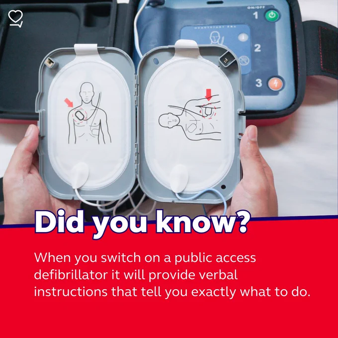Hands holding an open public access defibrillator displaying illustrated instructions on how to use it, with accompanying text explaining that the device provides verbal guidance when activated.