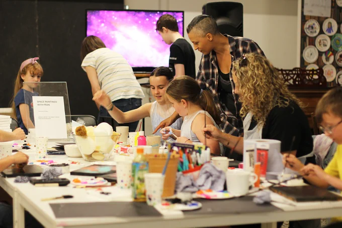 Artist Fran Williams working with a group of children on space paintings. Painting materials are spread across the table and a screen in the background plays images of space.