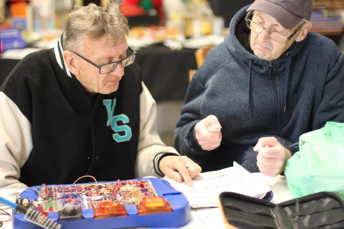 Two members of our men's creative wellbeing group are working on an electronics problem. They are sitting at a table with a book open and an appliance with a circuit board.