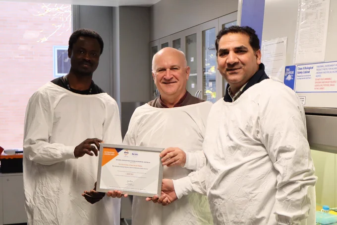 Photo of George Enninful, Mark Willcox and Muhammad Yasir holding the NATA accreditation certificate dressed in lab coats - photo taken in the PC laboratory 