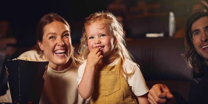 Two adults and a child enjoying a movie theater experience together, smiling and sharing popcorn.