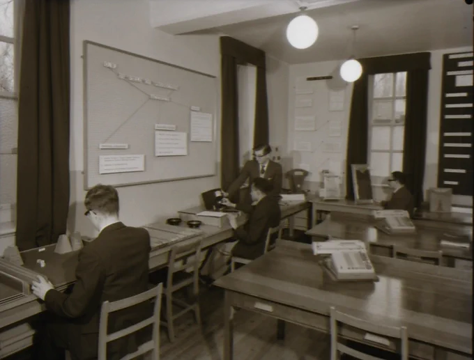 A black and white image. Four men are sitting at wooden desks in a lab. There is a board on the wall beside them and the windows have long curtains.