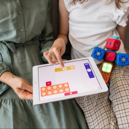 A person in a green dress sits next to a child in a white shirt and plaid pants. They are using a tablet with a colorful app displayed on it, featuring a grid and various icons. The child holds a stack of colorful, cube-shaped robots with lights and smiley faces, suggesting a playful, interactive learning activity.