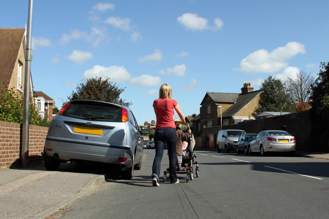 A blonde haired woman pushes a pram with a toddler and a baby in the middle of a residential road. There is a car on the pavement to the
left, blocking the path.