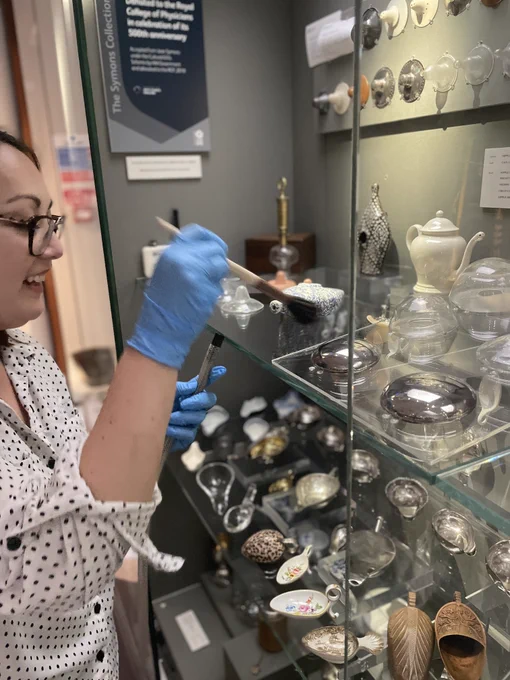 A woman in blue gloves uses a brush to dust around items in an open display case.