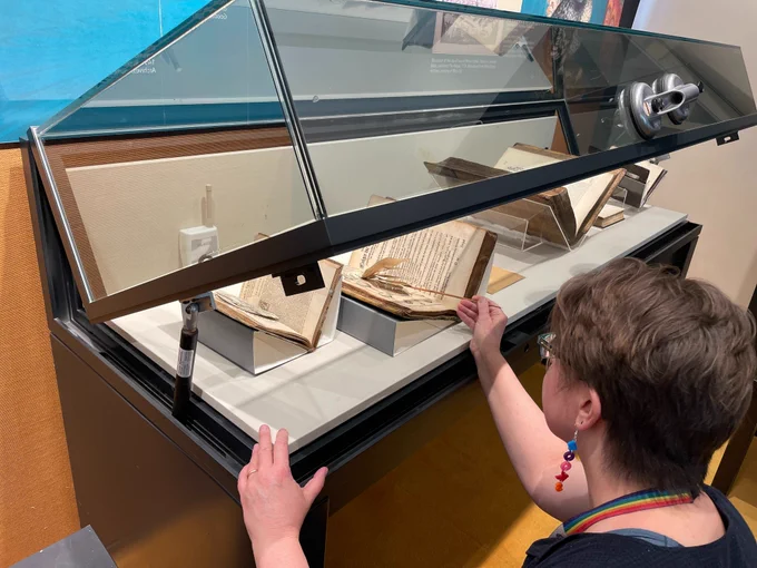 A woman crouches down and uses a stick to delicately lift the flaps of a diagram in a book inside a display case.