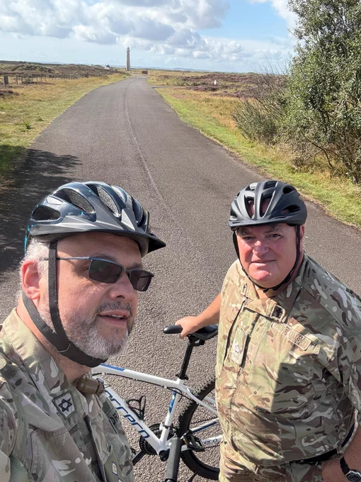 Two men in military terrain pattern uniforms wearing cycle helmets and holding pushbikes.  They are on a country road which leads to a lighthouse.