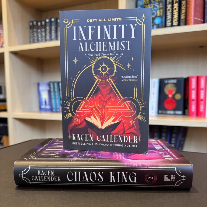 A copy of Infinity Alchemist by Kacen Callender standing on top of a copy of its sequel, Chaos King, in front of a bunch of bookshelves.