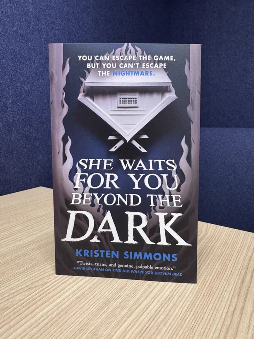 A copy of She Waits For You Beyond the Dark by Kristen Simmons standing on a table in front of a dark blue background.