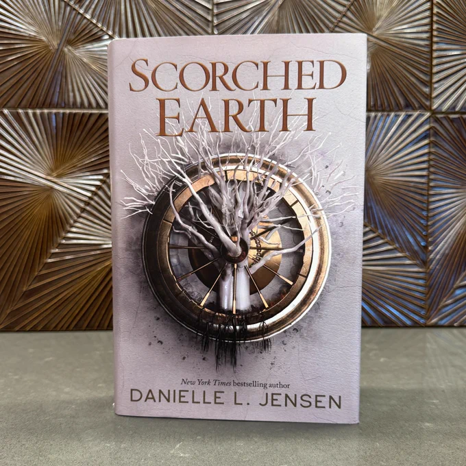 A copy of Scorched Earth by Danielle L. Jensen standing in front of a wall with bronze, patterned tiles.