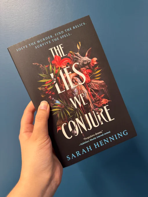 Someone holding up a coy of The Lies We Conjure by Sarah Henning in front of a blue wall.
