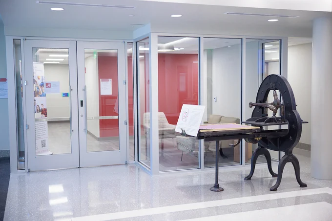 Interior space in the McLure Library. Featuring an antique printing press on display next to glass entrance doors. The hallway is well-lit, with informational posters on the walls and a visible lounge area through the doors.