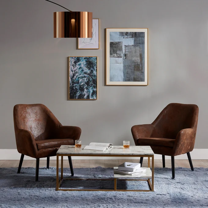 Two brown leather armchairs facing each other with a wooden coffee table in between, atop a gray rug. Above the table hangs a modern copper pendant light. The wall behind is adorned with three framed artworks.