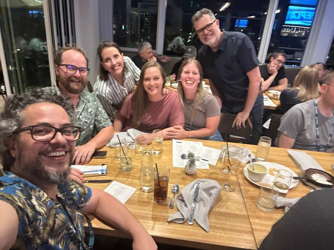 A group of six people smiling and posing around a wooden restaurant table with drinks and menus. The group looks cheerful, and the background shows other diners in a lively setting.