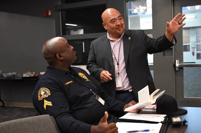 Man in suit gesturing while speaking to a male seated at a table during a group session.