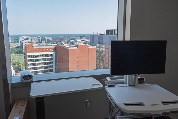 Office space with a desk and a monitor, overlooking a cityscape through a large window.