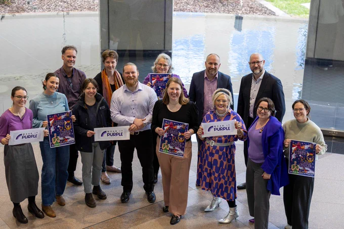ASNO staff standing in an atrium wearing purple and holding Wear it Purple day signs
