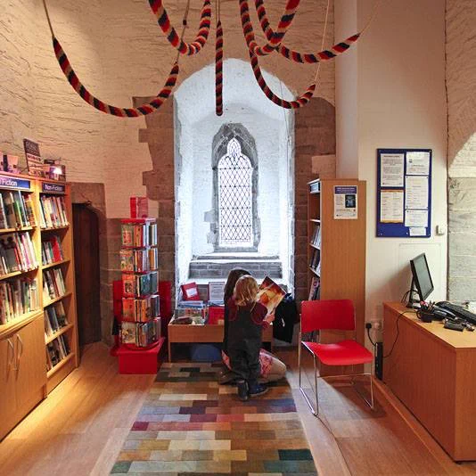 A cozy library set inside a historic stone building with arched windows. Colorful bell ropes hang from the ceiling, while shelves of books line the walls. Two children sit on a patterned rug reading together near a small display of books.