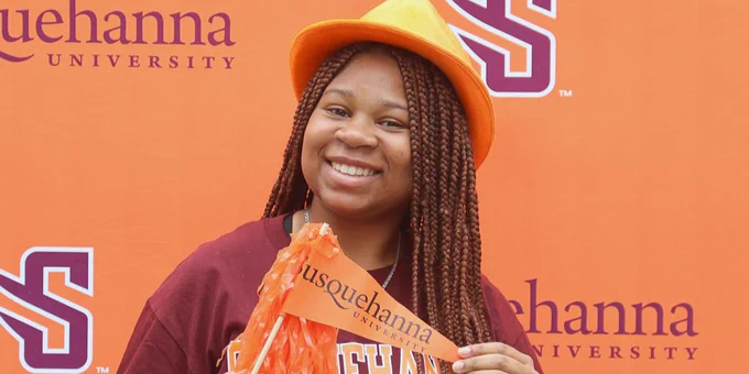 A student wears an orange hat and holds a pom-pom while standing in front of a backdrop featuring the Susquehanna University logo. She is smiling and dressed in university apparel.