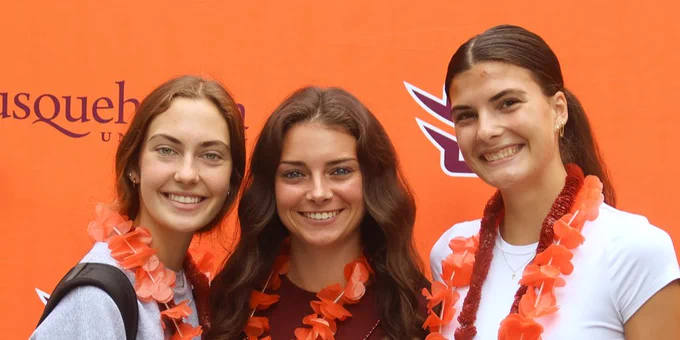 Three young women pose together, smiling in front of a vibrant orange backdrop with the logo of Susquehanna University. They each wear leis made of orange flowers, and one is dressed in a maroon soccer jersey. The atmosphere suggests a celebratory event or gathering.