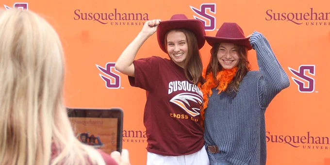 Two women pose happily in front of a Susquehanna University backdrop. One woman wears a maroon shirt and a matching hat, while the other sports an orange accessory and a gray sweater. A third person is taking their picture with a phone.