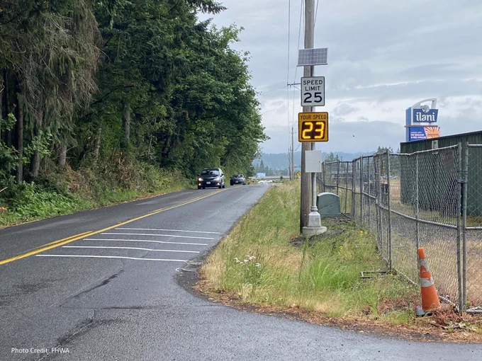 A car drives along a road with a 25 mph speed limit sign; a digital sign shows the current speed at 23 mph. A forested area and overcast sky are visible. 
