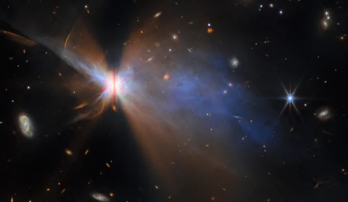 A wide-field image of IRAS 16594-4656 taken by the James Webb Space Telescope. The nebula’s bright core is split by a narrow dark band, with expansive rainbow lobes of light and color radiating outward. Numerous background galaxies and stars are visible across the field.