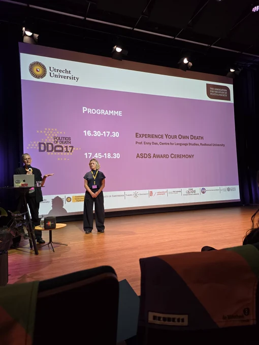 A conference stage features a large presentation screen displaying the logo of Utrecht University and the title “POLITICS OF DEATH DD17.” The programme outlines two events: “Experience Your Own Death” by Prof. Enny Das from Radboud University, scheduled from 16:30 to 17:30, and an ASDS Award Ceremony from 17:45 to 18:30. Two individuals, one gesturing while talking and the other standing with hands folded, are present on stage. The lower part of the image shows the backs of audience seats, some with colorful covers. Various university and organizational logos are lined up along the bottom of the presentation slide.