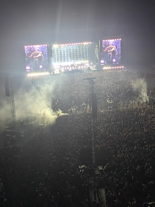 Nighttime stadium concert with a huge crowd. The band System of a Down is on stage, shown on two large screens as the members hug each other under bright lights and smoke.