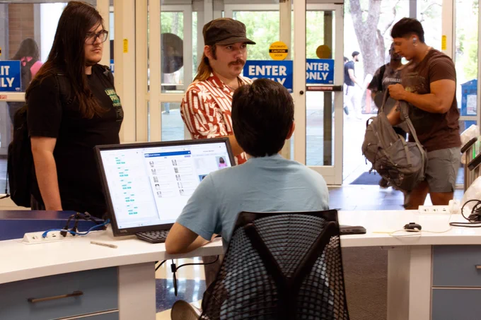 [ID: Pictured is a photo of a UTA Libraries staff member helping two students at the Welcome Desk on the first floor of the Central Library.]
