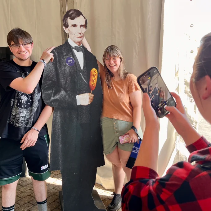 Two smiling fairgoers pose with a life-size cutout of Abraham Lincoln holding a corndog. One person points at “Corn Dog Abe” while another stands beside him. A third person takes their picture on a smartphone inside the Governor’s Tent at the DuQuoin State Fair.