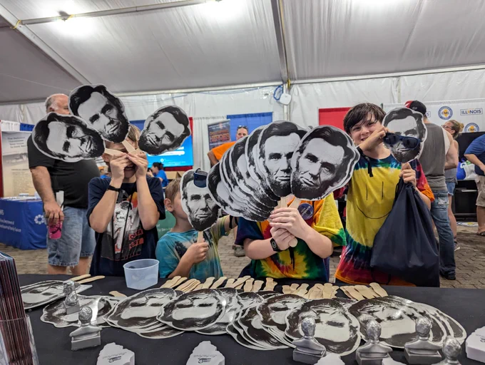 Children at the DuQuoin State Fair hold up Abraham Lincoln face fans in front of their faces while standing at a table covered with stacks of fans and Abe bust giveaways inside the Governor’s Tent.