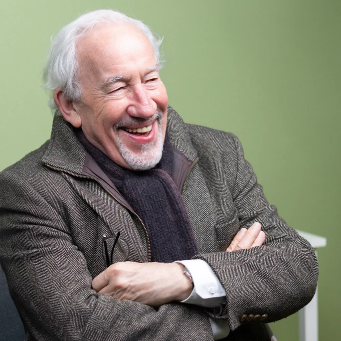 Simon Callow, pictured at the recording of Doctor Who: The Ingenious Gentlemen. He is seated, smiling, with his arms folder and his glasses in hand. 