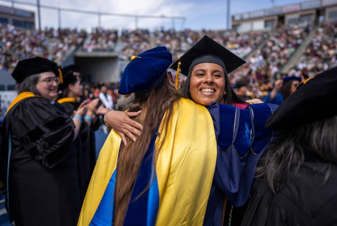 Smiling graduate in a cap and gown hugs a faculty member in academic regalia during a commencement ceremony.