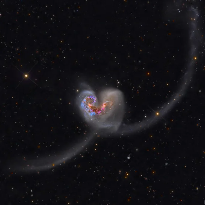 A deep-space image of the Antennae Galaxies, two colliding galaxies with long, sweeping tidal tails extending far into space. The galactic cores glow brightly with blue and red hues, indicating regions of intense star formation. Faint, wispy trails of stars and gas arc across a dark backdrop speckled with distant galaxies and stars.