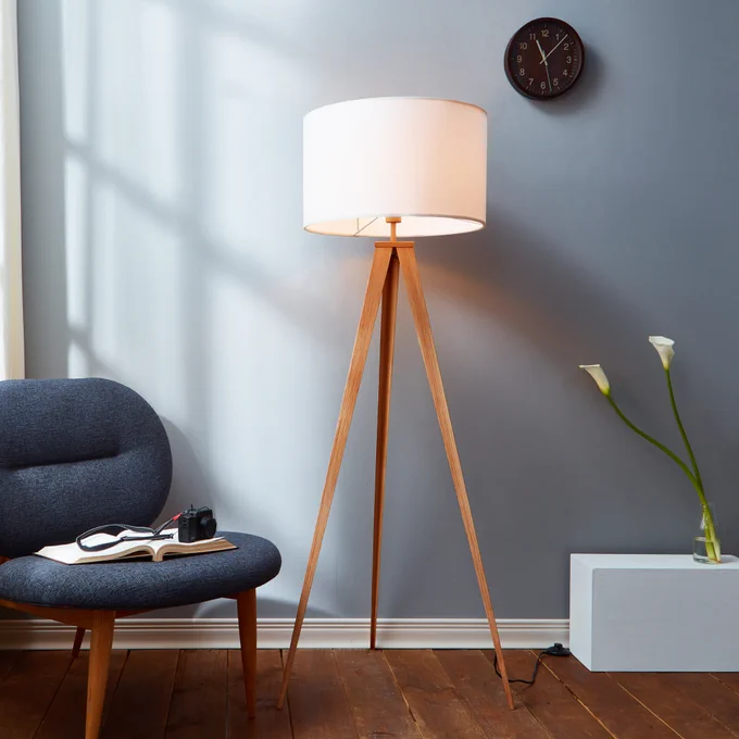 A modern living room corner featuring a stylish tripod floor lamp next to a comfortable blue armchair, with a white side table holding books and a camera. A clock hangs on the grey wall above.
