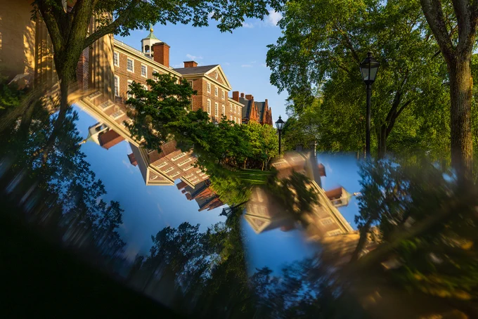 Panoramic view of University Hall with classic red brick buildings, trees, and lampposts, creatively reflected through clear glass revealing a blue sky.