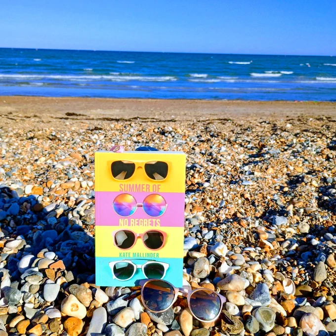 Copy of Summer of No Regrets by Kate Mallinder on a pebble beach with the sea in the background and a pair of sunglasses in the foreground