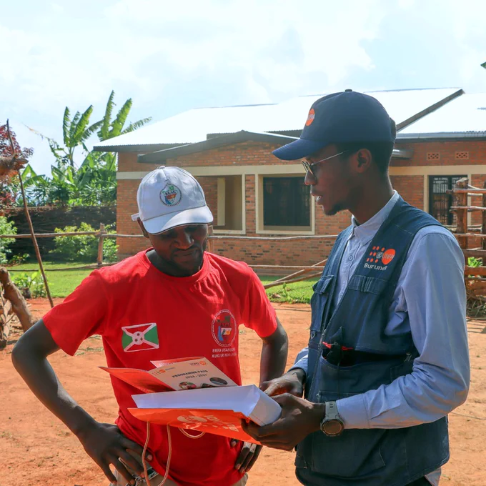 Two men stand outdoors near a brick building, one wearing a red shirt with a Burundi flag patch and the other in a blue vest holding documents.