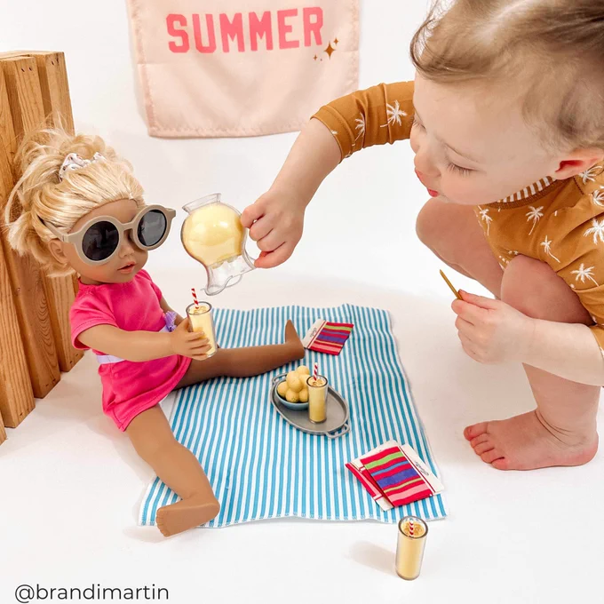 A child plays with a doll on a summer-themed setup, including a small picnic mat, beach accessories, and toy food items. The doll is wearing sunglasses and sitting casually.