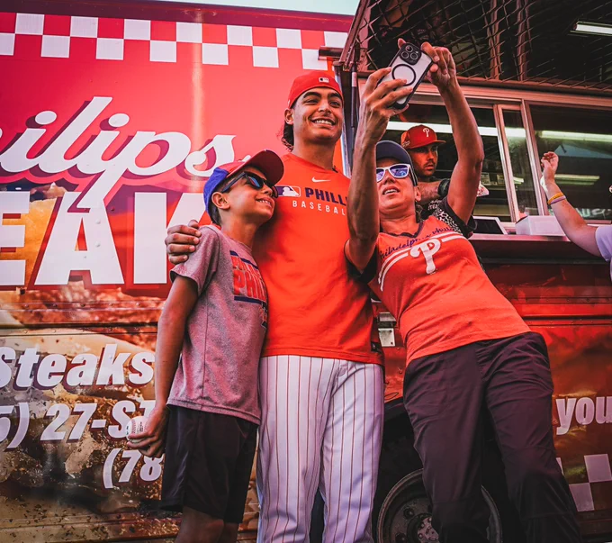 Jesús Luzardo smiles with fans taking a selfie at his Cheesesteak for Charity event. He's wearing the red phillies uniform and pinstripe pants.