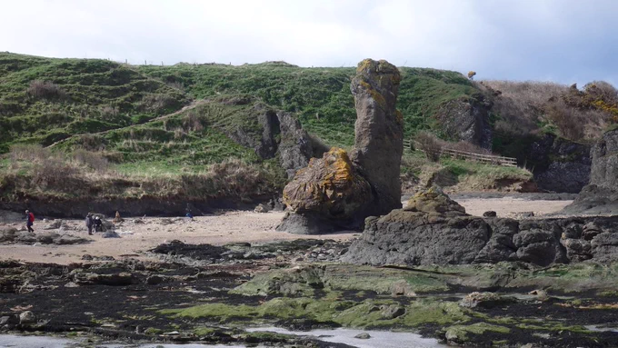 A rocky shore with grass covered cliffs in the background. On the sandy foreshore is a large rocky protrusion with a spire (spindle) and a round "rock" with radiating basalt columns, resembling a wagon wheel. 