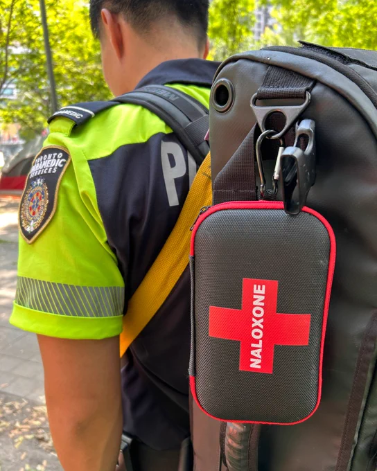 Community Paramedic in a bright yellow and navy uniform with a Toronto Paramedic Services patch, carrying a black backpack with a red and black naloxone kit attached.