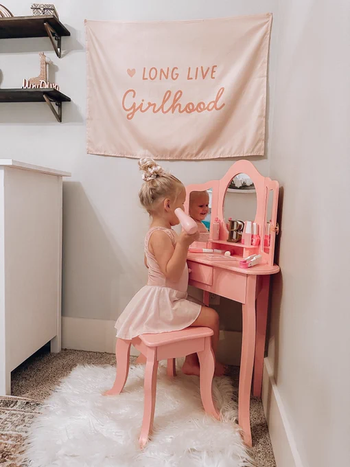 Child sitting at a pink vanity table, looking into a mirror and holding a brush, with a banner that reads "LONG LIVE Girlhood" hanging on the wall above.