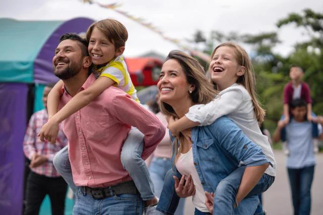 Group of four people smiling joyfully, with two adults each giving a piggyback ride to a child, at an outdoor event with a colorful tent in the background.