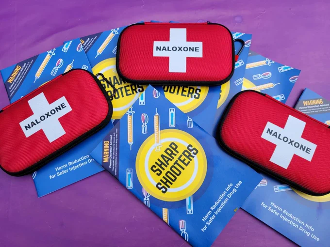 Three red naloxone kits with white crosses placed on blue harm reduction pamphlets titled 'Sharp Shooters'.