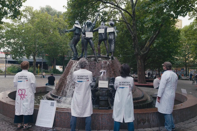 Aktivist:innen von Scientist Rebellion vor einem Brunnen, an dem Statuen die Augen verbunden wurden und Schilder mit "Ausbeutung", "Konsum" etc umgehängt wurden