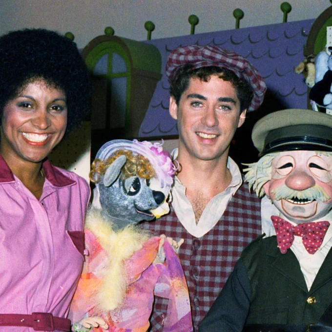 A group photo featuring three individuals and two puppets in a colorful, whimsical setting. The person on the left is a woman with a large afro hairstyle, wearing a pink dress with red trim and a matching belt, smiling warmly. The person in the center is a man with curly hair, wearing a red and white checkered vest and cap, also smiling. The person on the right is a man dressed as a puppet character with a white beard, large nose, and a colorful polka-dot bow tie, holding a puppet with a dog-like face adorned with a pink flower and yellow feathers. In the background, there are green and purple structures with arched windows and a display of various stuffed animal toys hanging on the wall.