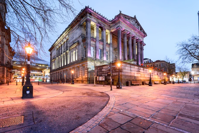Exterior of The Harris Museum Art Gallery during twilight hours. The building is a Free Public Library situated within a classical Grade I-listed museum building in Preston, Lancashire UK. On the pedestrian street in front of the building and on the road to its left, several street lamps are lit.